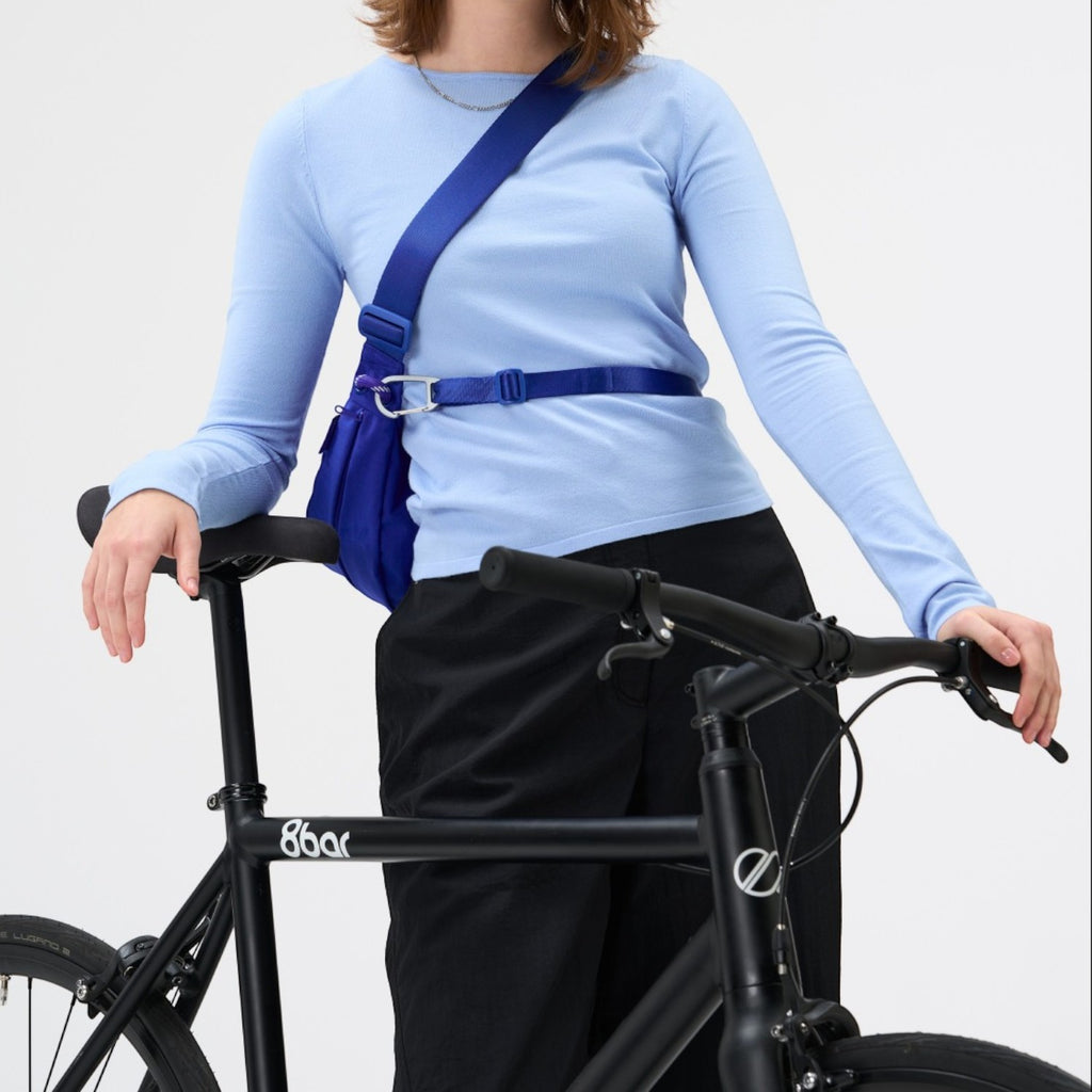 Woman wearing a bright blue Lieblingstasche sling bag across her chest while leaning on a black bicycle