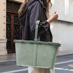 Lieblingstasche Carrybag in sage green with a sturdy aluminium handle, carried by a woman on a city street.