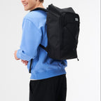 A man wearing a black Lieblingstasche Day Pack FLX backpack with a front bungee cord system on a white background.