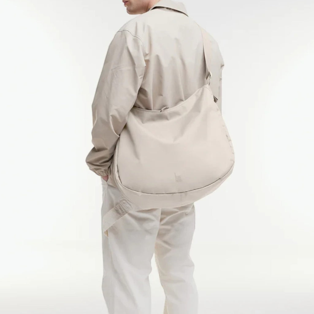 Lieblingstasche GOT BAG beige curved crossbody bag worn by a man in a matching light outfit against a white background