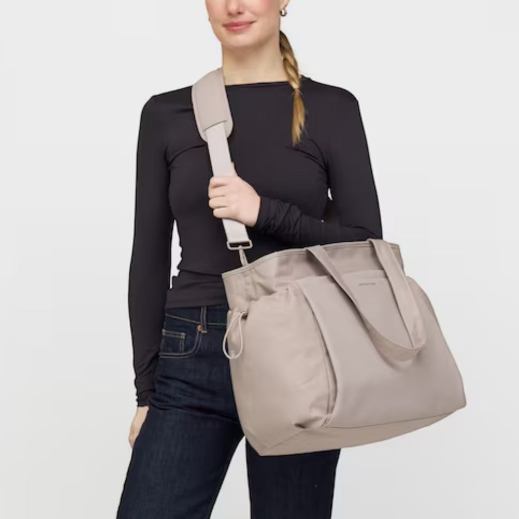 A woman wearing a beige Lieblingstasche Hellvi Pro Large tote bag with a wide shoulder strap against a white background.