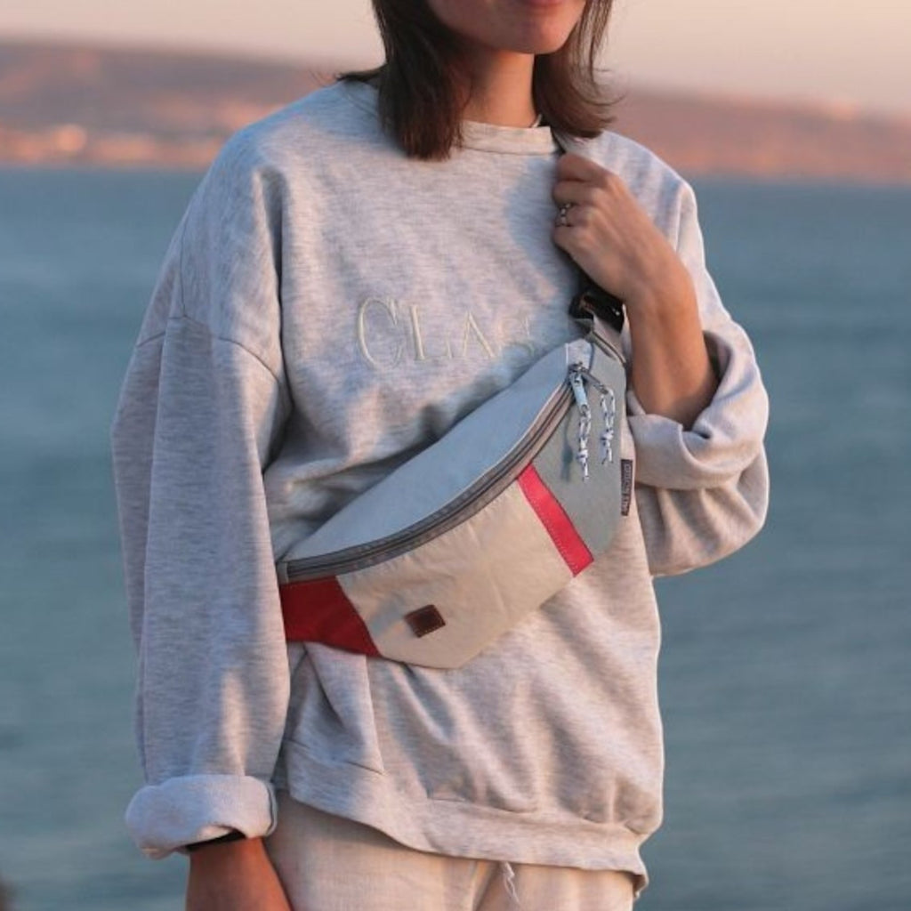 Lieblingstasche Knoten belt bag made from recycled white sailcloth with red accents worn by a woman at the coast.