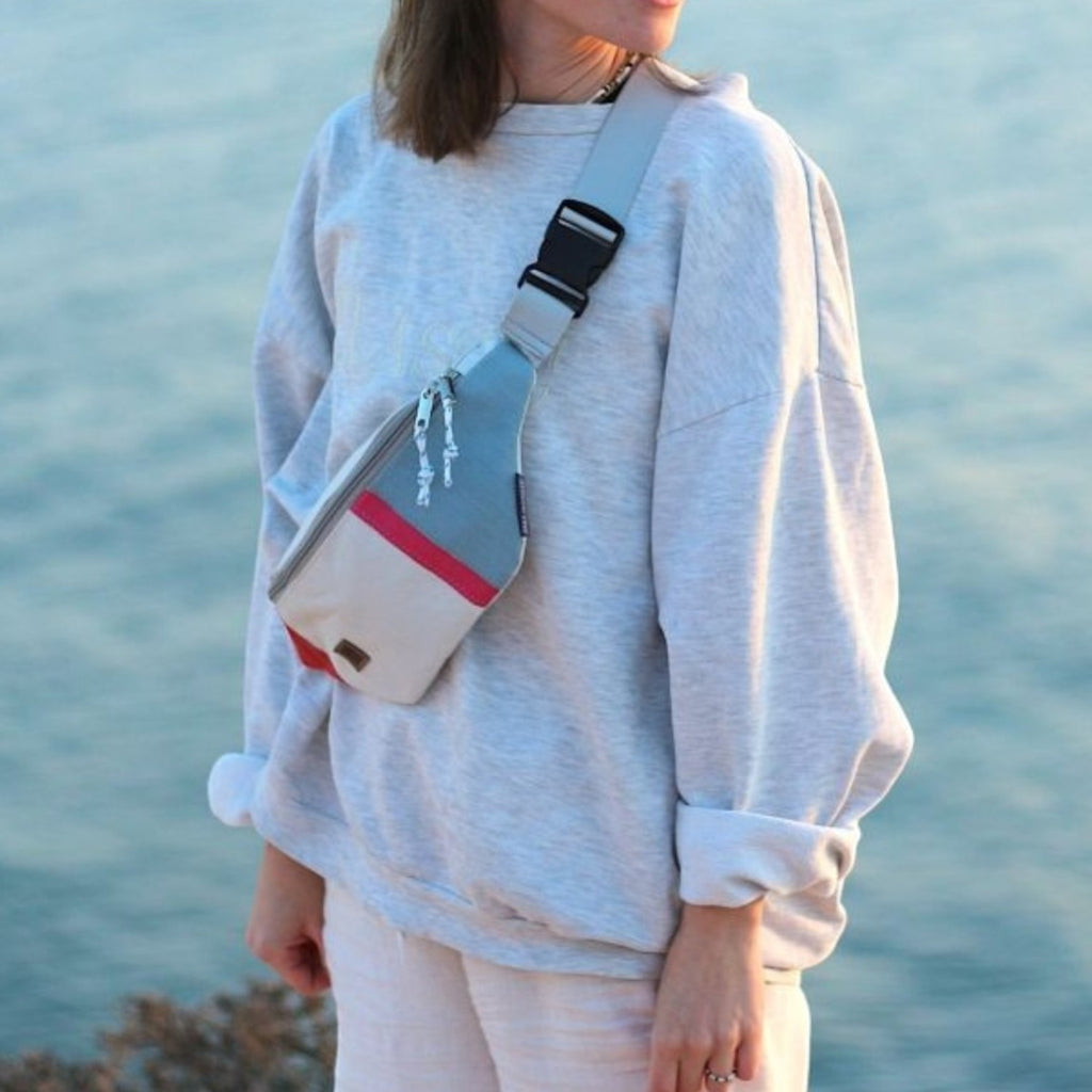 Lieblingstasche Knoten belt bag made from recycled sailcloth with a red accent, worn crossbody by a woman by the sea.