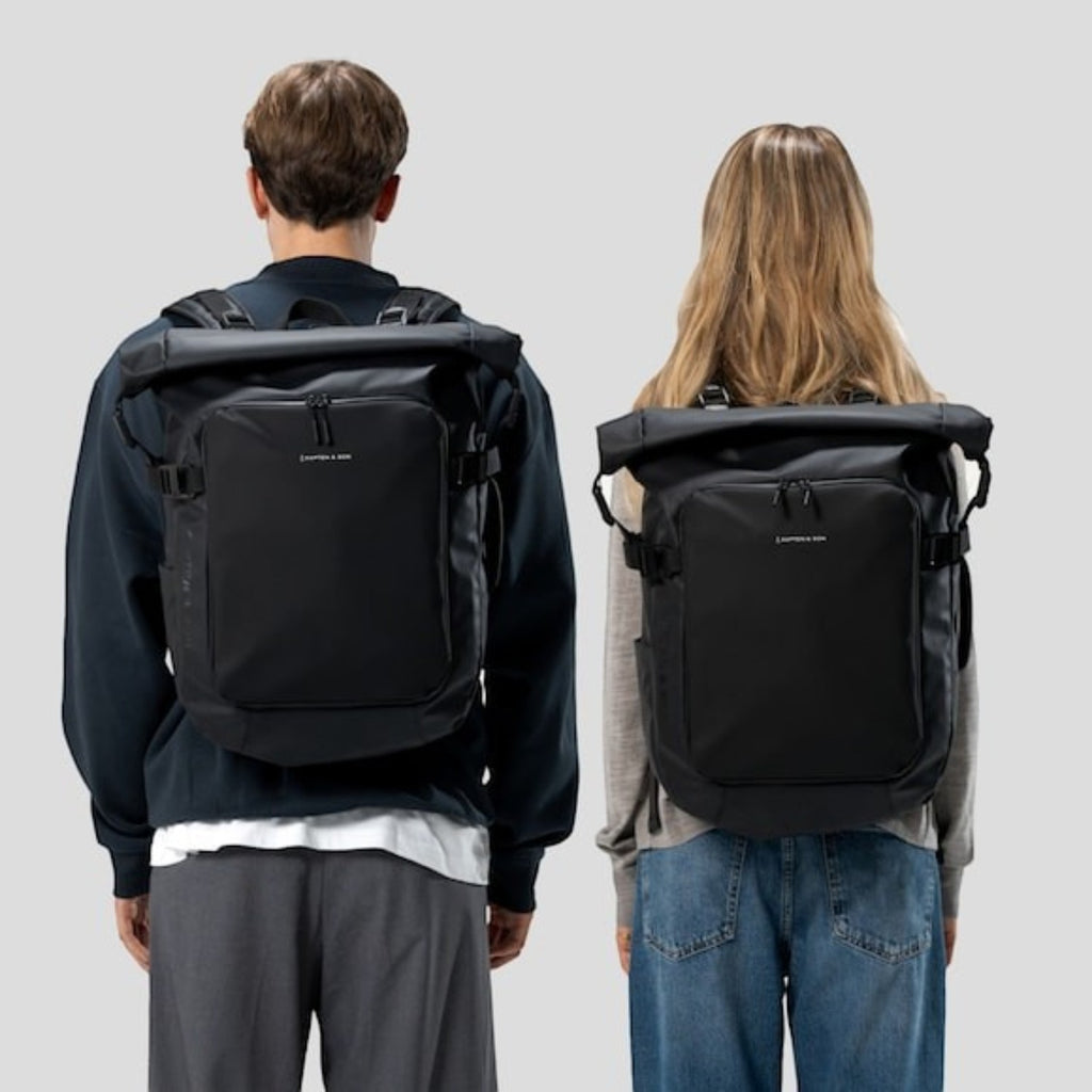 Lieblingstasche Lisbon Pro black rolltop travel backpack worn by a man and a woman against a light grey background.
