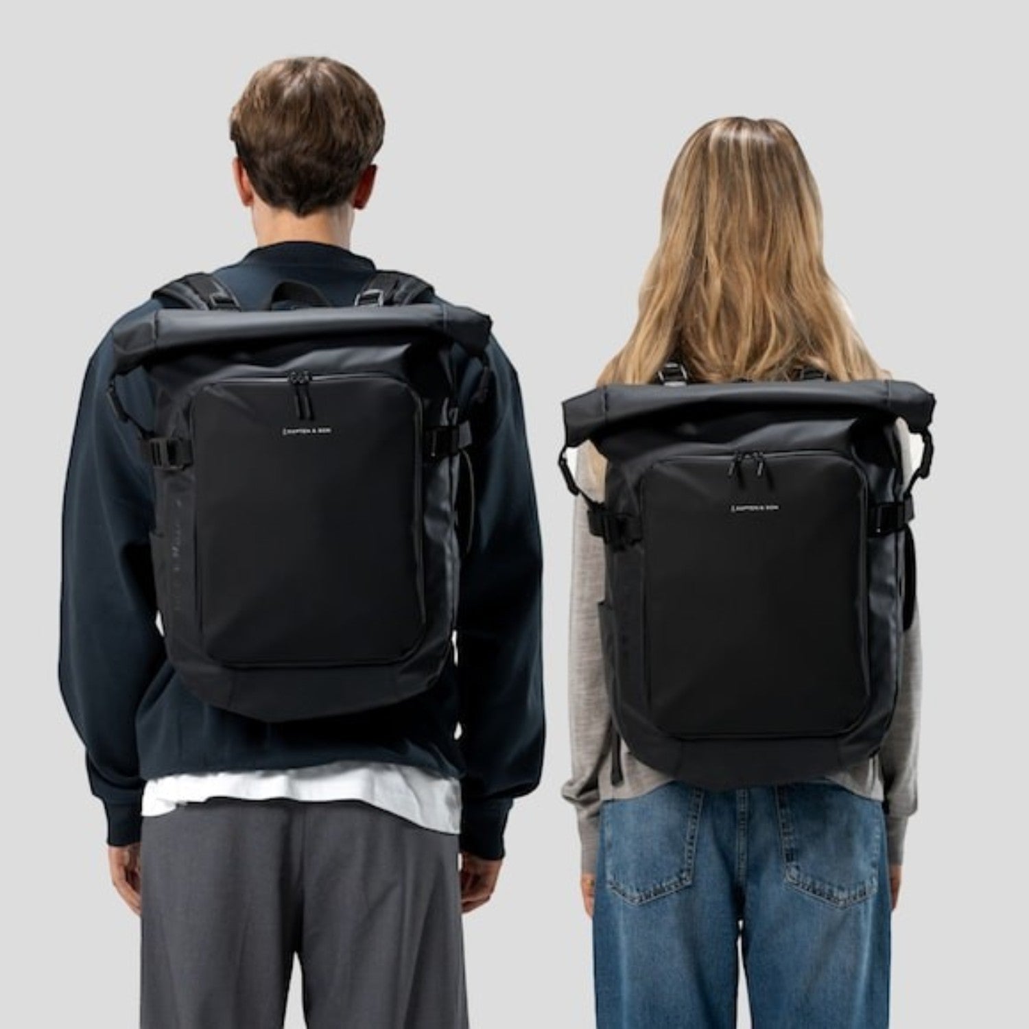 Lieblingstasche Lisbon Pro black rolltop travel backpack worn by a man and a woman against a light grey background.