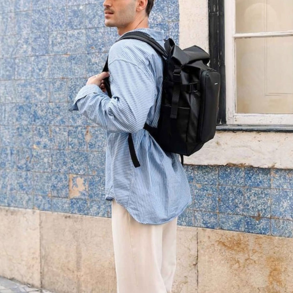 Lieblingstasche Lisbon small black backpack worn by a man in a blue striped shirt and cap against a blue tiled wall.