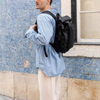 Lieblingstasche Lisbon small black backpack worn by a man in a blue striped shirt and cap against a blue tiled wall.