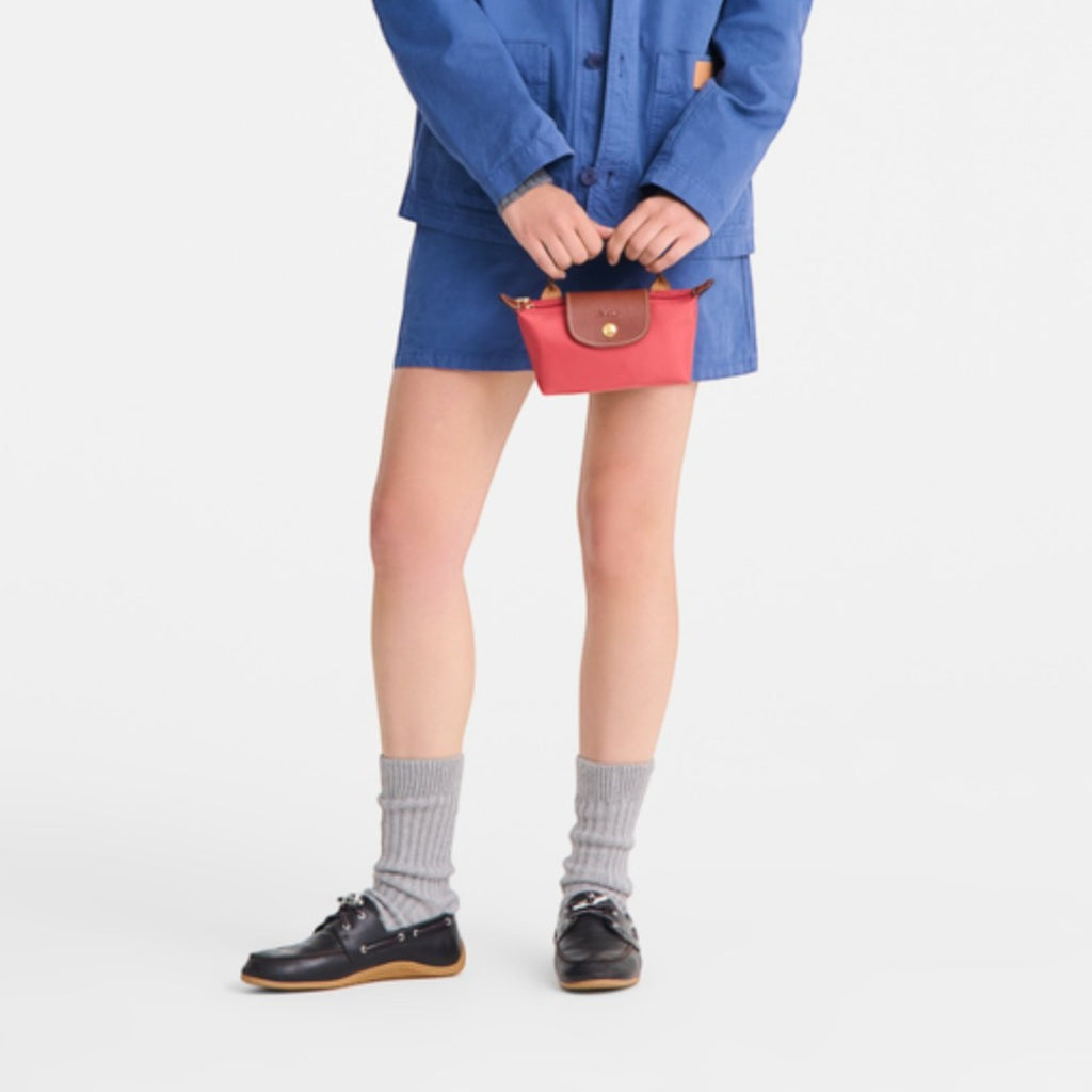 Model holding a small red Longchamp Le Pliage Pochette with brown leather trim, wearing a blue jacket and skirt.