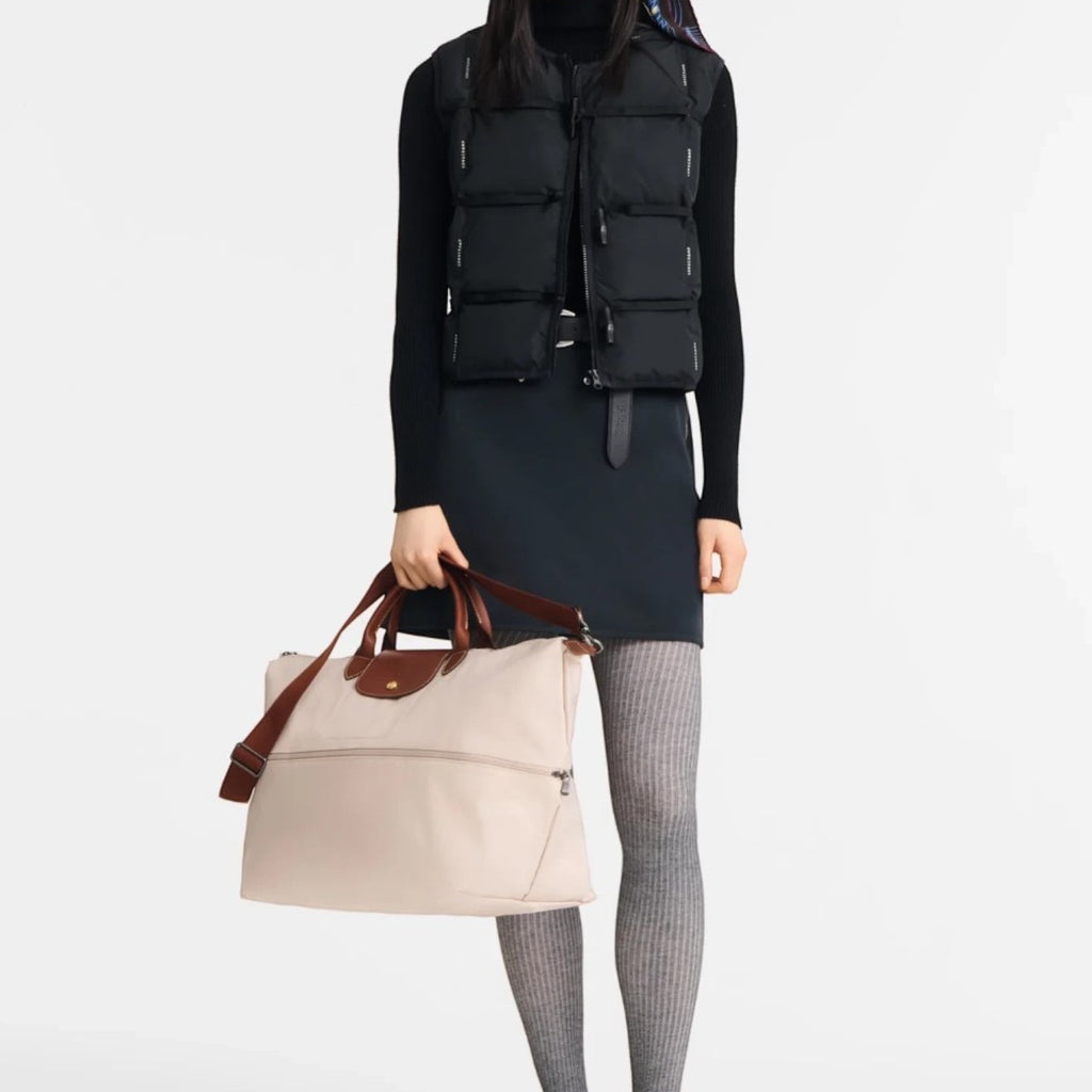 A model from Lieblingstasche wearing a black vest and holding a beige Longchamp Le Pliage travel bag with brown trim.