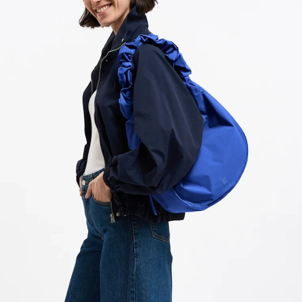 Lieblingstasche Moon Bag Ruffle Large in bright blue, worn as a shoulder bag by a smiling woman in a dark jacket.