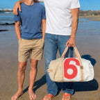 Lieblingstasche Pirat duffel bag with red number 6 on white canvas, held by a man on a sunny beach with a teenager.