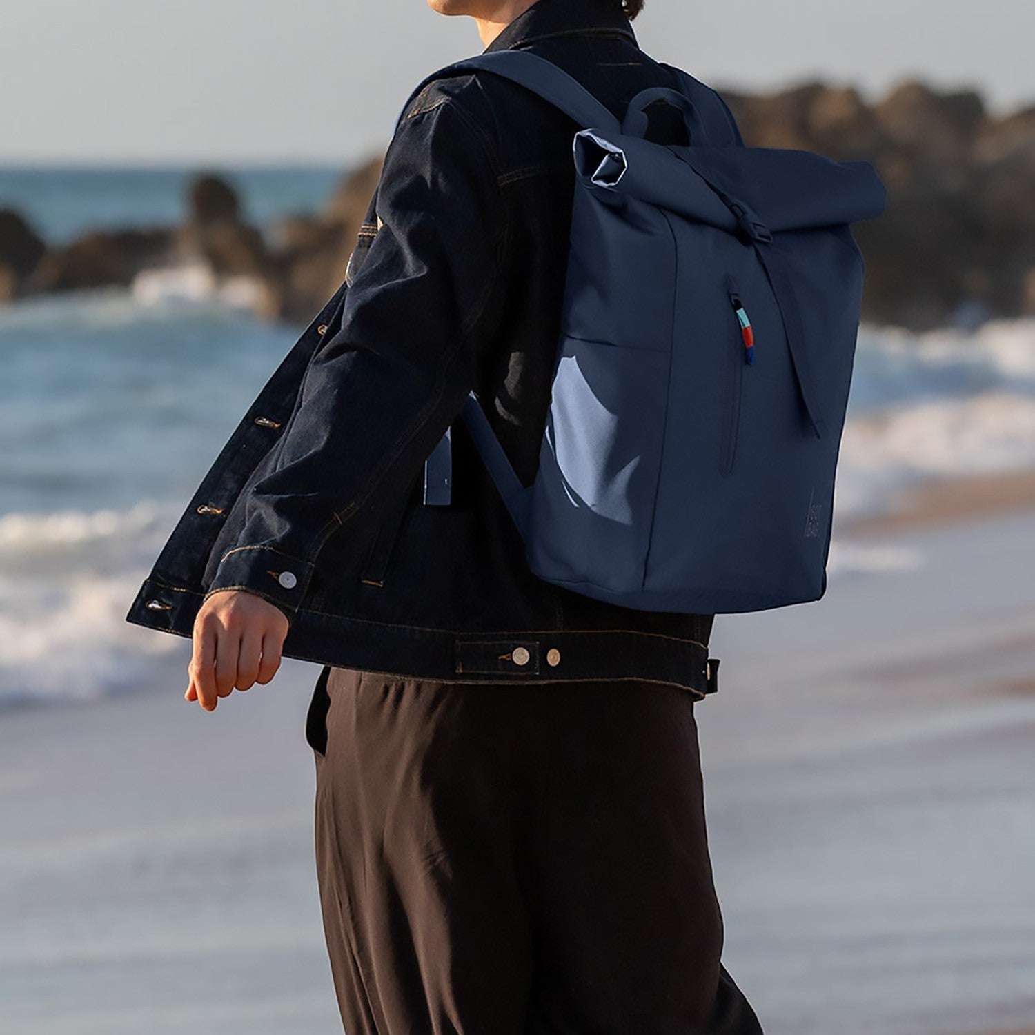 Lieblingstasche Rolltop Easy navy blue backpack with a roll-top closure and colorful zipper pull, worn by a person on a beach.