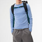 A man wearing a black Lieblingstasche Trip Pack backpack with green and blue straps over a light blue long-sleeve shirt