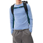 A man wearing a black Lieblingstasche Trip Pack backpack with green and blue straps over a light blue long-sleeve shirt